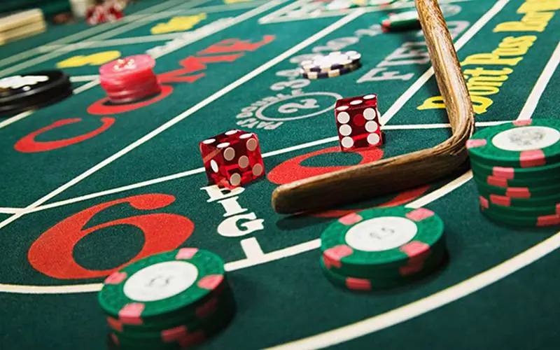 Dua dadu merah di meja craps hijau di samping tumpukan chip kasino hijau dan merah muda serta tongkat kayu bandar