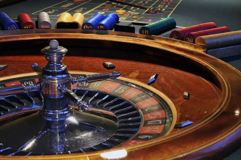 Close-up of a wooden roulette wheel with a silver turret, with rows of coloured casino chips on the betting table behind it
