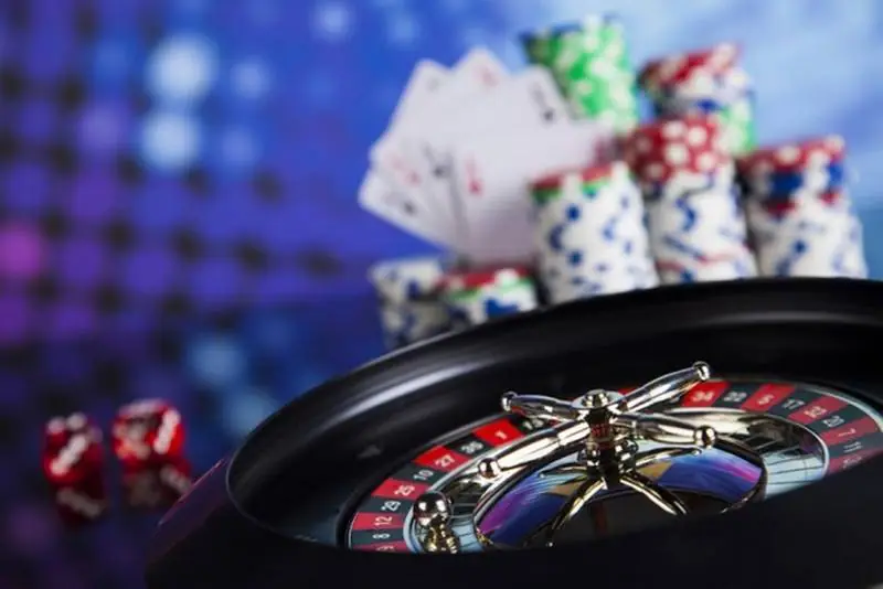 Roulette wheel in the foreground with stacks of casino chips, fanned playing cards and two red dice blurred behind it